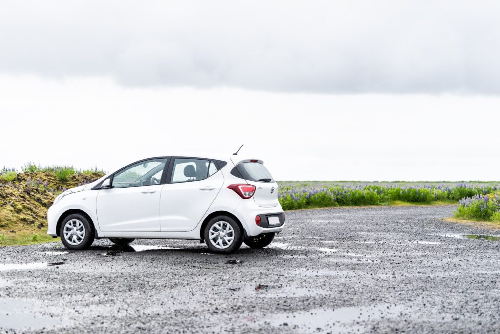 Small white Hyundai i10 smart car parked in parking lot by ring road with many lupine flowers, wet rain weather puddle, clouds
